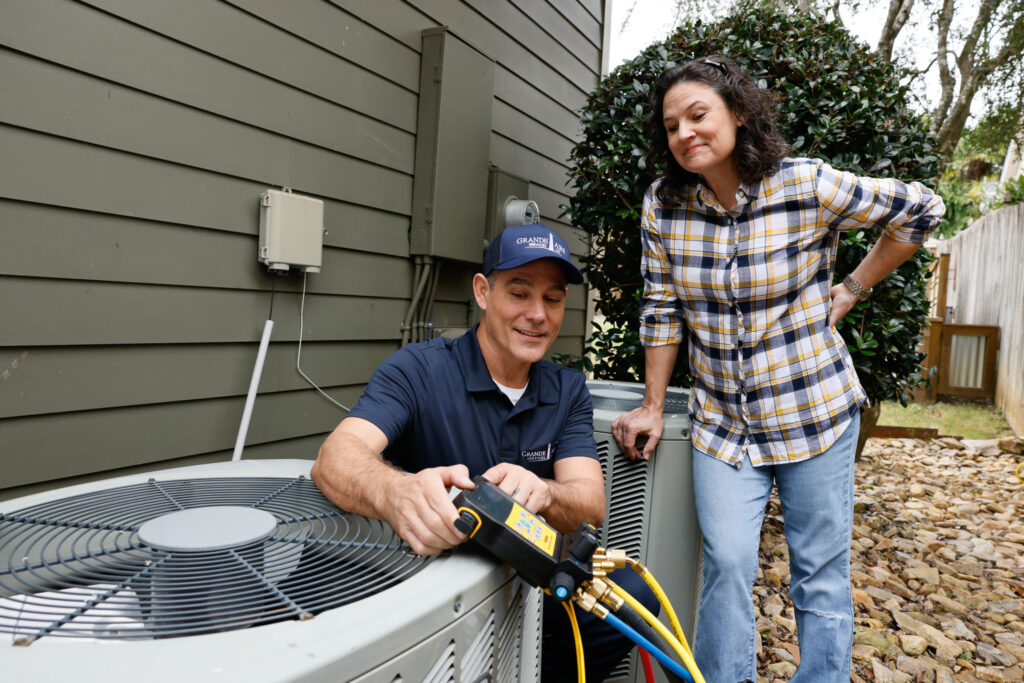 Homeowner watching a technician maintain an outdoor HVAC unit next to a gray/green home.