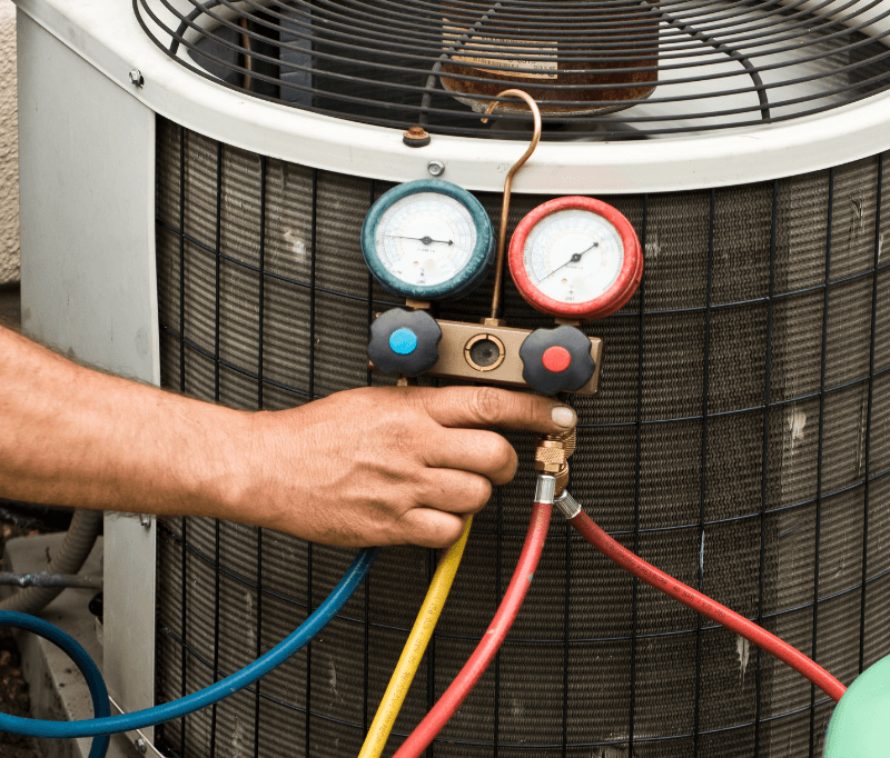 Technician's hand holding a gauge/meter in front of an outdoor HVAC unit.